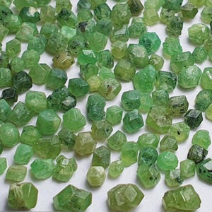 Könnte beinhalten: Eine Ansammlung kleiner, facettierter grüner Edelsteine, möglicherweise Peridot, auf einer weißen Oberfläche. Ein Lineal mit Zentimetermarkierungen befindet sich am unteren Rand, um den Maßstab anzugeben. Die Edelsteine variieren in Größe und Farbton.