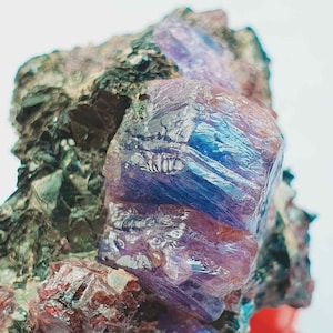 May include: A close-up of a raw, uncut gemstone with a striking display of colors. The gem features a prominent, crystalline structure with shades of deep purple and blue, set against a matrix of dark green and reddish-brown minerals. The gem is displayed on a red stand.