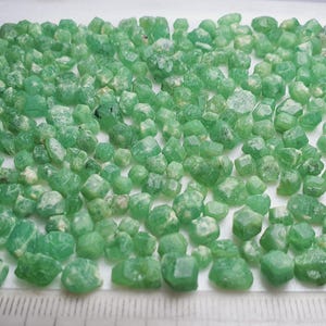 May include: A collection of small, faceted green gemstones, possibly emeralds, are scattered across a white tray. A ruler with inch markings is placed along the bottom, providing scale. The gems have a translucent quality.