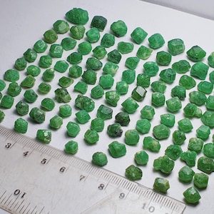 Può includere: Una collezione di piccole gemme verdi sfaccettate, forse smeraldi o minerali simili, disposte su una superficie bianca. Un righello con misure in centimetri è posto vicino alle gemme. Le gemme variano per dimensioni e tonalità di verde.