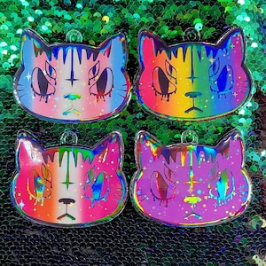Op de afbeelding: Vier regenboogkleurige holografische katten sleutelhangers met een druppelend verf effect. Elke sleutelhanger heeft een ander kleurenschema: blauw, oranje, roze en paars. De katten hebben een zwart kruis op hun voorhoofd en witte ogen.
