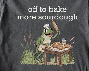Op de afbeelding: Donkergrijs t-shirt met de tekst "off to bake more sourdough". De afbeelding toont een cartoon kikker met een koksmuts en schort, die brood bakt. Het woord "pepper" staat onderaan.