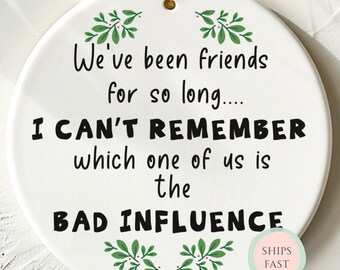 May include: A white ceramic ornament with a gold string for hanging. The ornament features the text "We've been friends for so long... I CAN'T REMEMBER which one of us is the BAD INFLUENCE" in black, with green leaf accents.