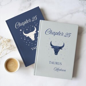 May include: Two hardcover books with the text "Chapter 25" and a Taurus bull illustration. One book is navy blue, the other is light gray. The navy book has a white bull illustration, and the gray book has a navy bull illustration. The word "Taurus" is below the bull.