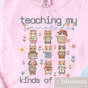 Puede incluir: Camiseta rosa claro con la frase "teaching my favorites kinds of blossom". El diseño presenta nueve gatos de dibujos animados vestidos de profesores, rodeados de flores. El texto y el gráfico son de estilo lúdico.