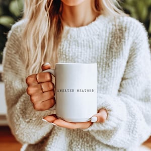 May include: A white ceramic mug with the text "SWEATER WEATHER" in black lettering. The mug is held by a person wearing a white sweater. The mug has a large handle and a cylindrical shape, perfect for enjoying warm beverages.