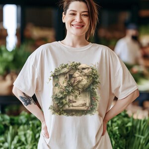 May include: A cream-colored short-sleeve t-shirt featuring a graphic print of a brown owl perched inside a weathered wooden birdhouse covered in green ivy leaves. The design is centered on the chest of the shirt, showcasing detailed textures of the wood, feathers, and foliage in muted, natural tones.