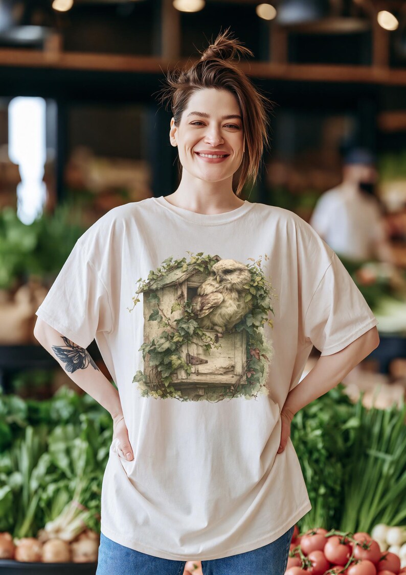 May include: A cream-colored short-sleeve t-shirt featuring a graphic print of a brown owl perched inside a weathered wooden birdhouse covered in green ivy leaves. The design is centered on the chest of the shirt, showcasing detailed textures of the wood, feathers, and foliage in muted, natural tones.