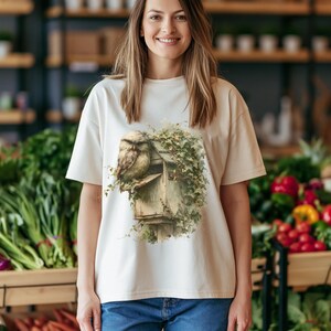 May include: A cream-colored short-sleeve t-shirt featuring a graphic print of a brown owl perched on a weathered wooden mailbox covered in green ivy vines. The shirt has a crew neckline and a relaxed fit, paired with blue denim jeans.