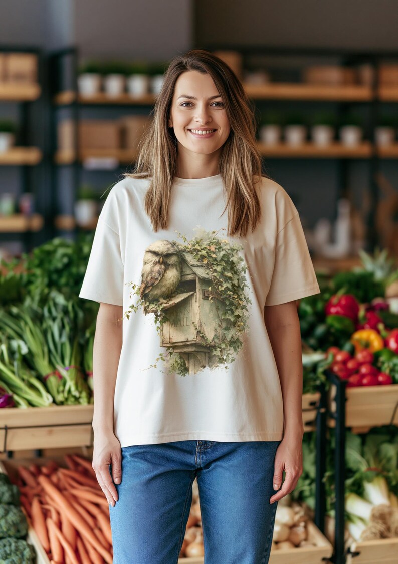 May include: A cream-colored short-sleeve t-shirt featuring a graphic print of a brown owl perched on a weathered wooden mailbox covered in green ivy vines. The shirt has a crew neckline and a relaxed fit, paired with blue denim jeans.