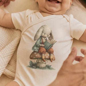 May include: A cream-colored short-sleeve baby bodysuit featuring a central graphic illustration of a rabbit wearing a light blue jacket and brown pants, sitting on a spotted mushroom while reading an open book with green covers.