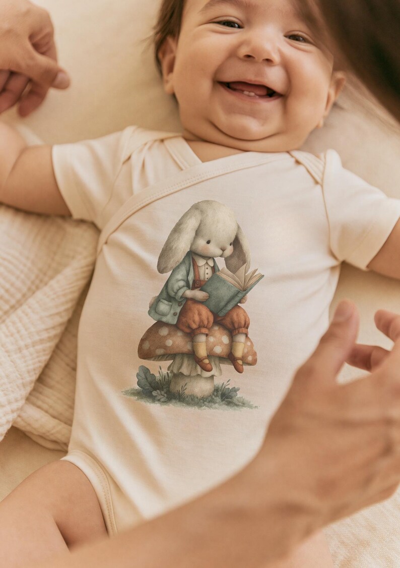 May include: A cream-colored short-sleeve baby bodysuit featuring a central graphic illustration of a rabbit wearing a light blue jacket and brown pants, sitting on a spotted mushroom while reading an open book with green covers.