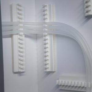 May include: White plastic tubing is routed through a series of white plastic clips mounted on a white wall. The tubing is curved and forms a loop.