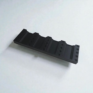 Puede incluir: Un objeto rectangular negro con múltiples agujeros y crestas. El objeto, sobre un fondo blanco, parece estar hecho de un material sólido. El diseño sugiere un componente mecánico o industrial.