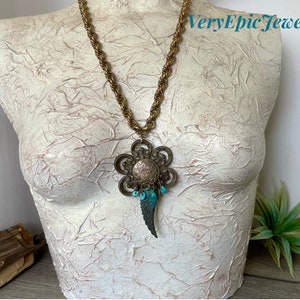 May include: A gold chain necklace with a large, ornate pendant featuring a flower design with turquoise accents. The pendant also has a wing-shaped charm.