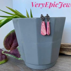 Op de afbeelding: Roze en witte gemarmerde druppelvormige oorbellen met zilverkleurige bloemaccenten. De oorbellen worden getoond op een grijze geometrische plantenbak met groene en paarse bladeren. De tekst "VeryEpic Jewelry" is zichtbaar.