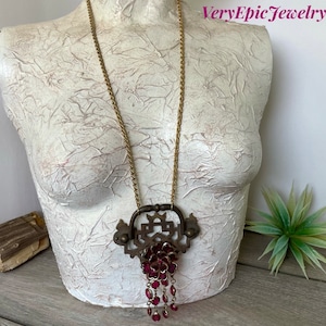 May include: A gold chain necklace with a large, ornate pendant featuring a geometric design and a cluster of dangling burgundy red beads. The pendant is made of brass and has a vintage look.