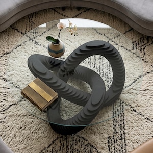 Op de afbeelding: Een moderne glazen salontafel met een unieke grijze knoopvormige voet. De tafel heeft een helder glazen blad, een kleine vaas met een bloem en een decoratief boek. De tekst onderaan luidt: DXF - AI - DWG - EPS SVG - PDF.