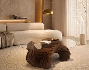 May include: A coffee table featuring a circular glass top resting on a dark brown, sculptural wooden base with layered, wave-like horizontal slats. The base has a fluid, organic shape that supports the transparent surface from below.