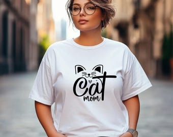 Puede incluir: Camiseta blanca con un gráfico de cara de gato negro y las palabras "Cat mom" en fuente cursiva. La camiseta tiene mangas cortas y cuello redondo.