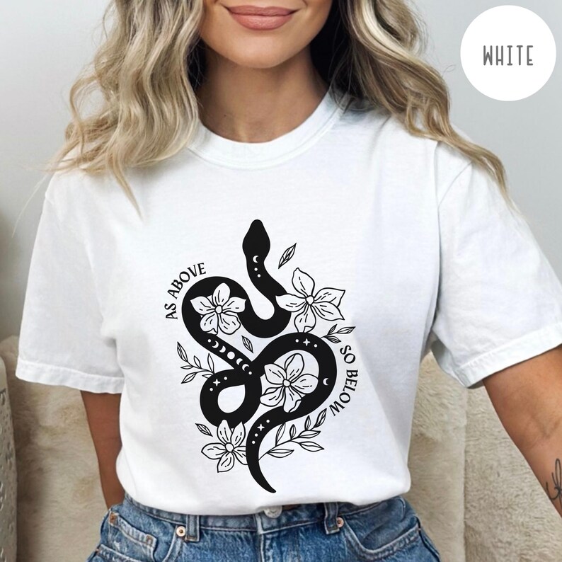 Pu&ograve; includere: T-shirt bianca con un disegno di serpente nero intrecciato con fiori, lune e stelle. Il testo "AS ABOVE SO BELOW" &egrave; arcuato attorno al serpente. Stile casual.