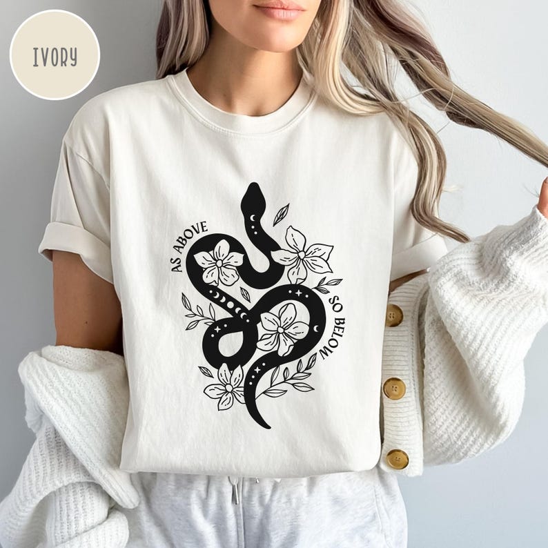 Pu&ograve; includere: T-shirt color avorio con un disegno di serpente nero intrecciato con fiori e la scritta "AS ABOVE SO BELOW". Il design include elementi floreali e celesti. La maglietta ha maniche corte e una vestibilit&agrave; ampia.
