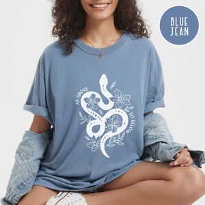 Pu&ograve; includere: Una maglietta blu jeans con una grafica bianca di un serpente intrecciato con fiori e la scritta "AS ABOVE SO BELOW". La maglietta ha maniche arrotolate e scollo tondo. Un cerchio blu con le parole "BLUE JEAN" &egrave; nell'angolo in alto a destra.