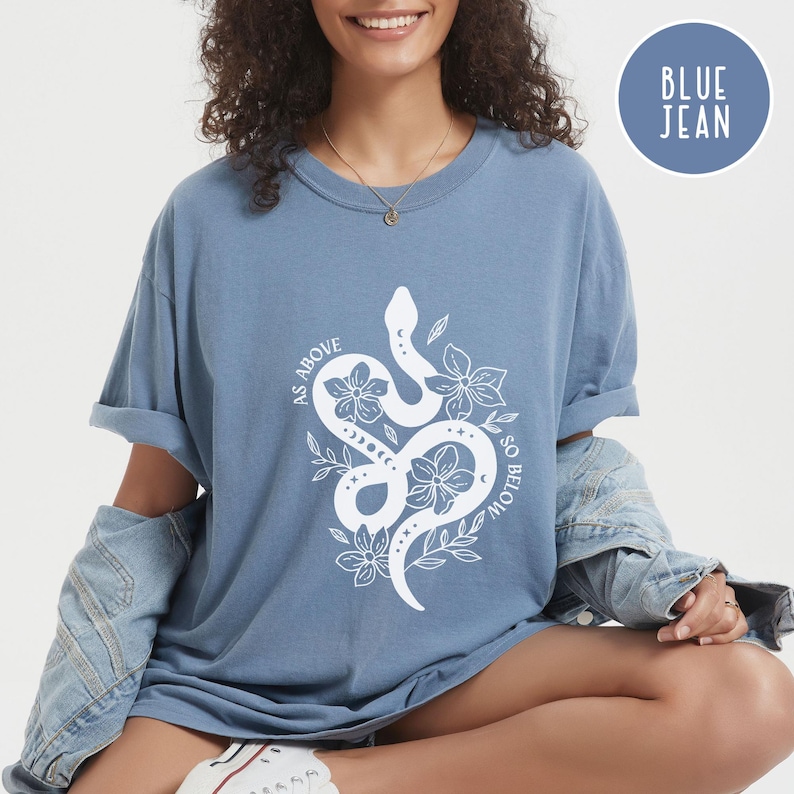Pu&ograve; includere: Una maglietta blu jeans con una grafica bianca di un serpente intrecciato con fiori e la scritta "AS ABOVE SO BELOW". La maglietta ha maniche arrotolate e scollo tondo. Un cerchio blu con le parole "BLUE JEAN" &egrave; nell'angolo in alto a destra.