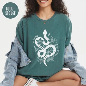 Pu&ograve; includere: T-shirt color verde acqua con un disegno di serpente e fiori bianchi. Il testo "AS ABOVE SO BELOW" &egrave; stampato intorno al serpente. La maglietta ha una vestibilit&agrave; ampia ed &egrave; realizzata in un materiale morbido. Il colore &egrave; "BLUE-SPRUCE".