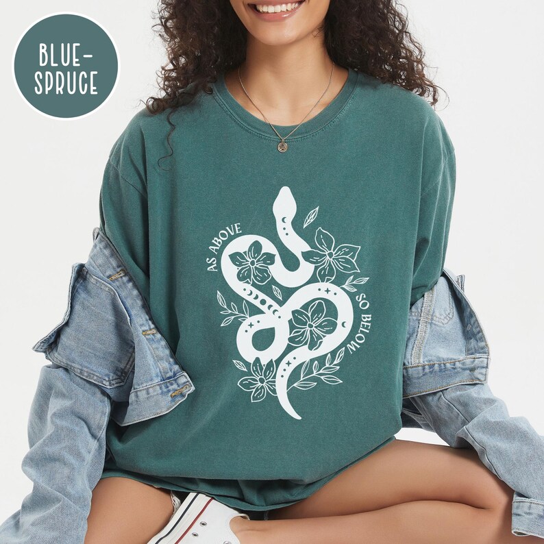 Pu&ograve; includere: T-shirt color verde acqua con un disegno di serpente e fiori bianchi. Il testo "AS ABOVE SO BELOW" &egrave; stampato intorno al serpente. La maglietta ha una vestibilit&agrave; ampia ed &egrave; realizzata in un materiale morbido. Il colore &egrave; "BLUE-SPRUCE".