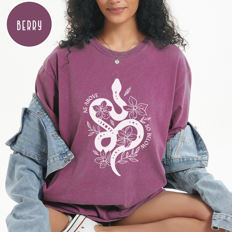 Pu&ograve; includere: T-shirt a maniche lunghe color bacca con grafica bianca di serpente e fiori. Il testo "AS ABOVE SO BELOW" &egrave; stampato intorno al disegno del serpente. La modella indossa una giacca di jeans.