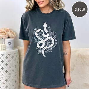 Pu&ograve; includere: T-shirt grigio scuro con una grafica bianca di un serpente intrecciato con fiori e la scritta "AS ABOVE SO BELOW". La maglietta ha maniche corte e girocollo. La maglietta &egrave; color Pepe.