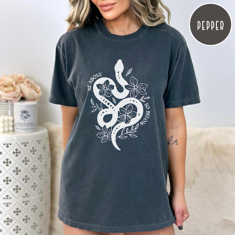 Pu&ograve; includere: T-shirt grigio scuro con una grafica bianca di un serpente intrecciato con fiori e la scritta "AS ABOVE SO BELOW". La maglietta ha maniche corte e girocollo. La maglietta &egrave; color Pepe.