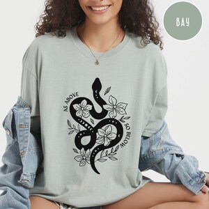 Pu&ograve; includere: T-shirt verde salvia con un design di serpente nero intrecciato con fiori e la scritta "AS ABOVE SO BELOW". Il design include fasi lunari ed elementi floreali. La maglietta ha una vestibilit&agrave; ampia.