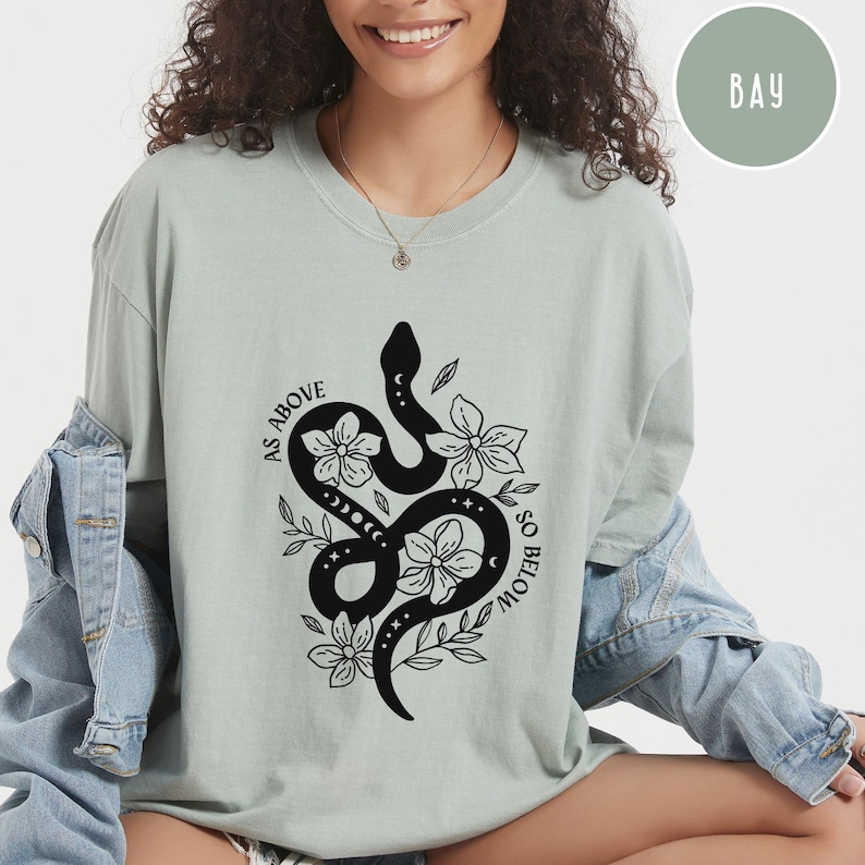 Pu&ograve; includere: T-shirt verde salvia con un design di serpente nero intrecciato con fiori e la scritta "AS ABOVE SO BELOW". Il design include fasi lunari ed elementi floreali. La maglietta ha una vestibilit&agrave; ampia.