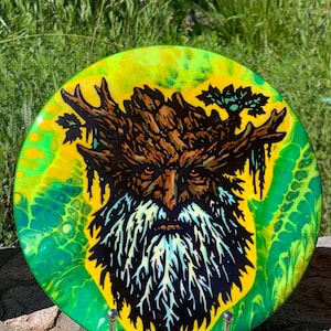 Pode incluir: Um disco circular com um fundo de arte fluida em amarelo e verde vibrantes. Ao centro, uma ilustração detalhada de uma figura barbuda com um rosto composto por casca de árvore, ramos e folhas, em tons de castanho, preto e branco.