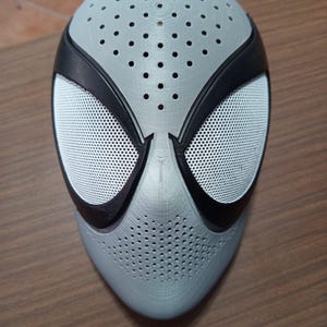 May include: A gray and black Spider-Man mask with white mesh eye coverings. The mask has numerous small holes across the surface and is made of a hard material. The mask is designed to cover the entire face.