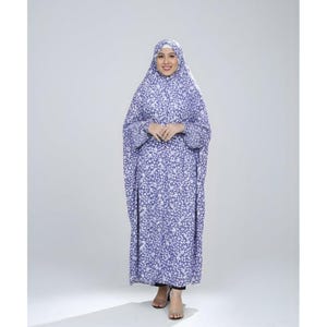 May include: A full-length, light purple prayer garment with a white floral pattern. The garment includes a head covering and long sleeves. The person is wearing black pants and sandals. The background is a plain white.