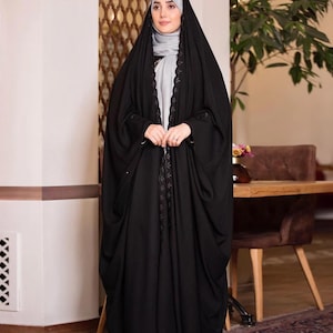 May include: A full-length image of a person wearing a black abaya. The abaya is a long, flowing garment with a decorative trim down the front. The person is also wearing a gray hijab. The background includes a wooden table and a plant.