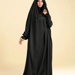 Può includere: Un'abaya nera a tinta unita con cappuccio drappeggiato, maniche lunghe con polsini elastici arricciati e un dettaglio a linguetta orizzontale sul petto. L'indumento è lungo fino ai piedi con una silhouette ampia e fluida che copre il corpo.