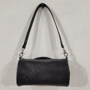 以下が含まれることがあります： 黒い革のハンドバッグ。長くて調節可能なストラップとシルバーカラーの金具が付いています。長方形のバッグには上部にハンドルとジッパー開閉部があります。白い壁にバッグが吊り下げられています。