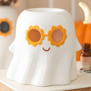 May include: A white ceramic ghost-shaped container with orange flower-shaped sunglasses and a smiling face. The container is part of a Halloween or autumn-themed collection. An orange pumpkin is in the background.
