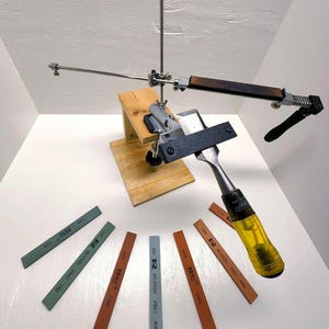 May include: A woodworking chisel sharpening system with a wooden base, metal components, and a black handle. Several sharpening stones in various colors and grits are arranged around the system.
