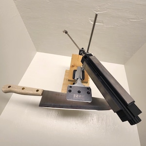 May include: A knife sharpening system with a large cleaver-style knife. The system includes a metal guide, a wooden base, and a sharpening stone. The knife has a wooden handle and a wide, rectangular blade.