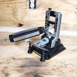 May include: A Work Sharp knife sharpener, featuring a black base, silver and gray components, and a sharpening stone. The device is designed to sharpen knives and tools. The image is set against a wooden background.