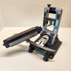 May include: A silver and black Work Sharp knife sharpener with a knife in place. The sharpener has a black base, a silver frame, and a sharpening guide. The Work Sharp logo is visible on the base.