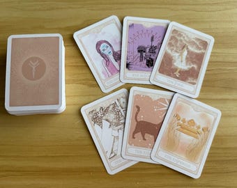 May include: A deck of tarot cards with a light brown back featuring a central botanical icon, alongside six face-up cards displaying illustrations of a person, a cat, a crown, and other figures in shades of pink, brown, and white with printed titles.
