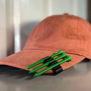 May include: A terracotta coloured baseball cap with a black golf tee holder attached to the brim. Four green golf tees are inserted into the holder.