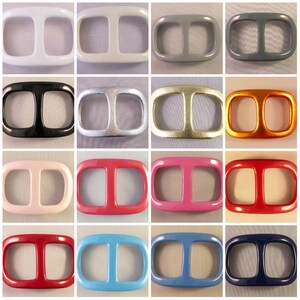 May include: A collection of sixteen rectangular belt buckles in various colors. The buckles have a rounded rectangular shape with two rectangular openings. Colors include white, gray, black, silver, gold, orange, pink, red, and blue.