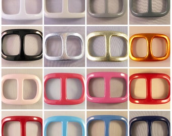 May include: A collection of sixteen rectangular belt buckles in various colors. The buckles have a rounded rectangular shape with two rectangular openings. Colors include white, gray, black, silver, gold, orange, pink, red, and blue.